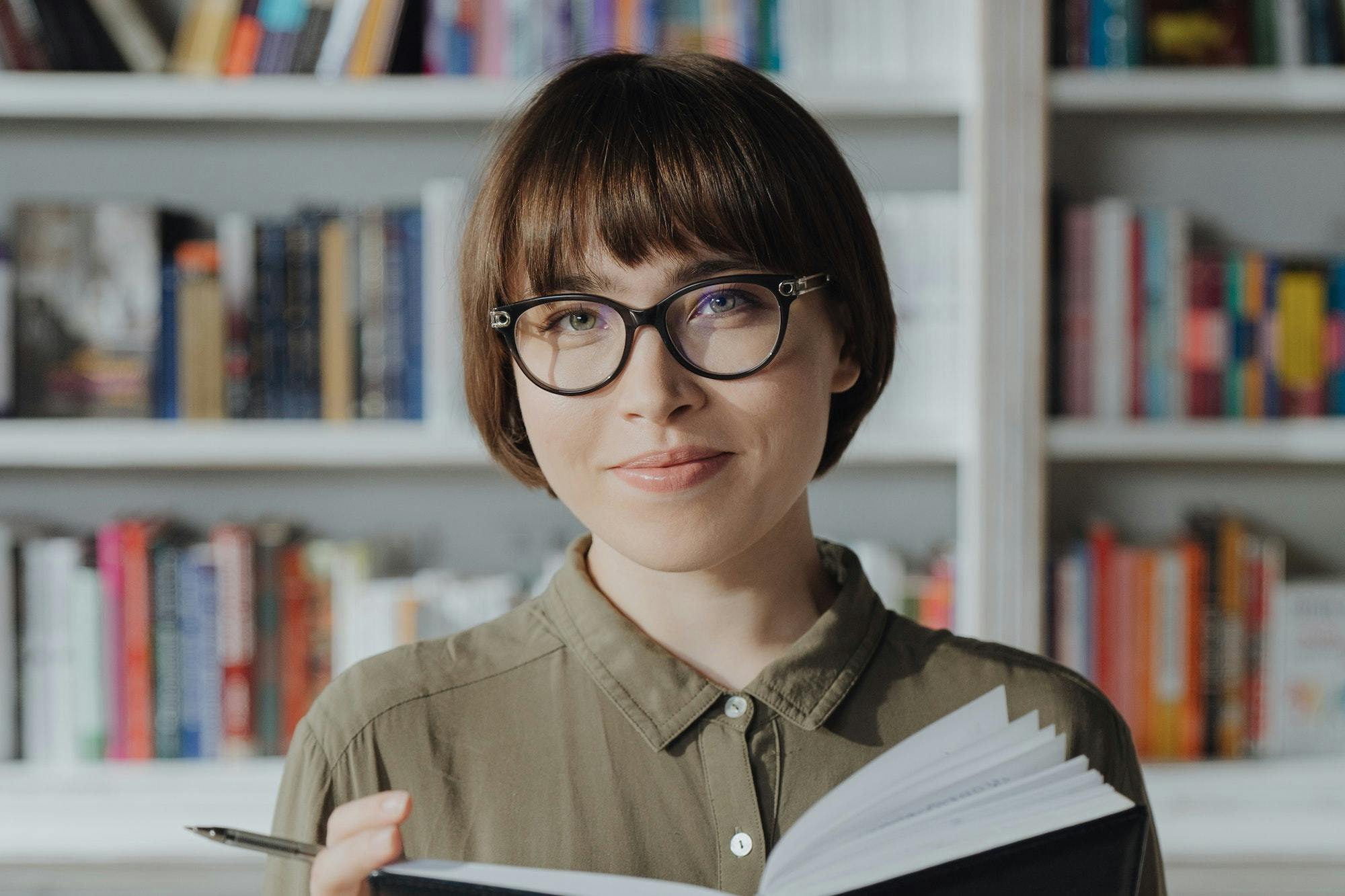 Junge Frau mit Brille und Buch vor Bücherregal.